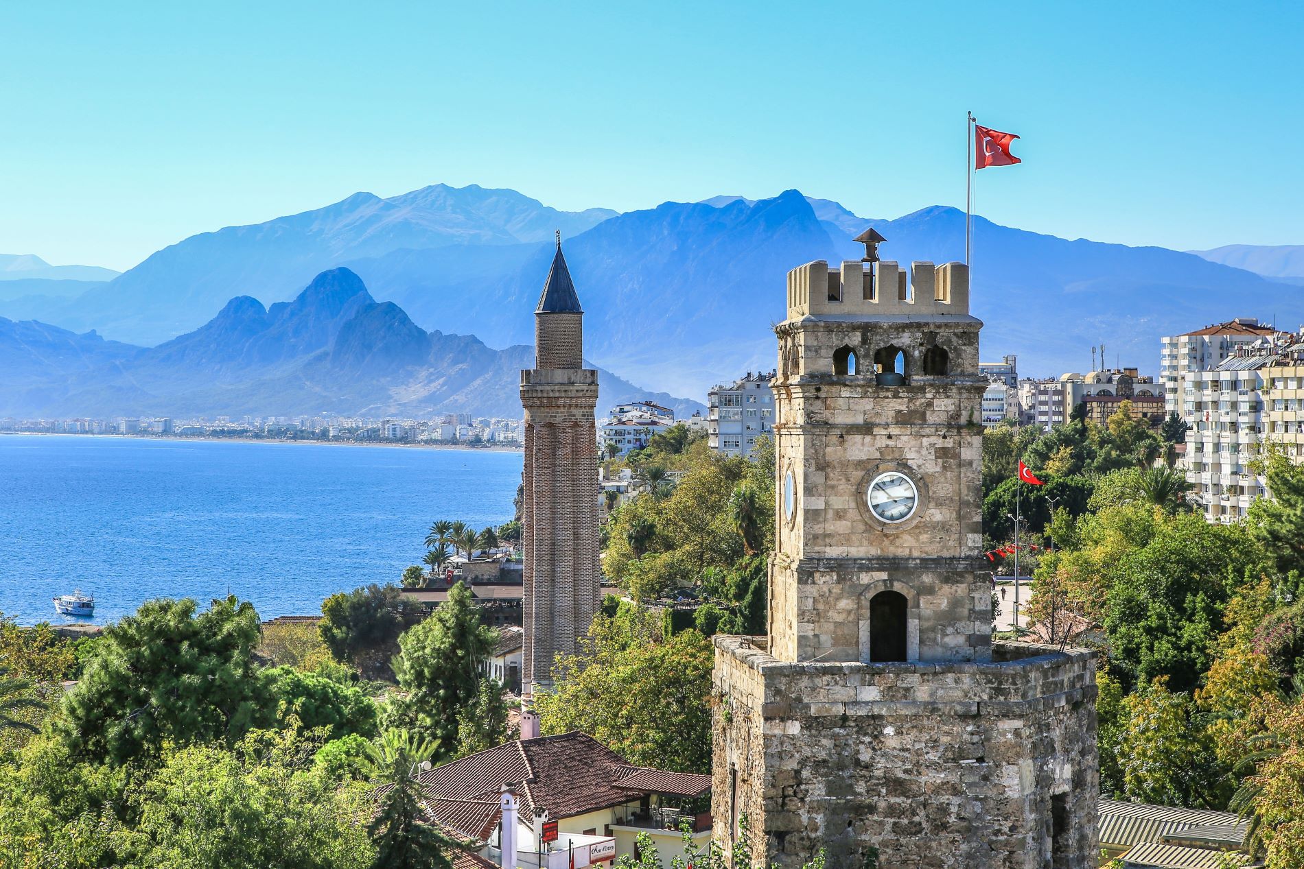 Bick auf den Uhrenturm Saat Kulesi im Vordergrund, der aus der  Römerzeit, stammt an einem sonnigen Sommertag. Im Hintergrund ist der Strand und die Stadt zu sehen. Am Horizont türmt sich das Gebirge auf.