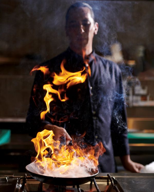 Mann bereitet Essen in einer Pfanne zu. Aus der schiesst eine hohe Flamme hervor.