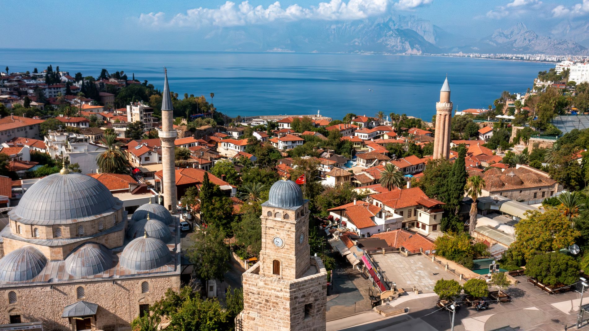 Bick auf den Uhrenturm Saat Kulesi im Vordergrund, der aus der  Römerzeit, stammt an einem sonnigen Sommertag. Im Hintergrund ist der Strand und die Stadt zu sehen. Am Horizont türmt sich das Gebirge auf.