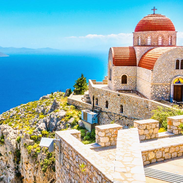 Ein aus hellem Stein gebautes Kloster mit markanten roten Kuppeln und Ziegeldächern, umgeben von karger Felsenlandschaft. Im Hintergrund erstreckt sich das weite, blaue Meer unter einem klaren Himmel.