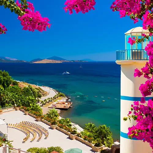 Ein weißer Leuchtturm mit blauen Streifen steht rechts, umgeben von leuchtend pinker Bougainvillea. Der Blick schweift über eine Hotelterrasse und eine Bucht mit klarem, blauem Wasser. Im Hintergrund liegen Inseln und Berge unter strahlend blauem Himmel.