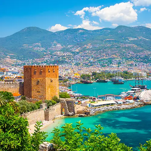 Der mittelalterliche Rote Turm (Kızıl Kule) aus rotem Stein dominiert den Hafen von Alanya in der Türkei. Türkisblaues Meer, ein belebter Hafen, Stadtbebauung und bewaldete Berge im Hintergrund.
