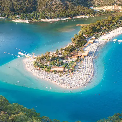 Ein sichelförmiger Sandstrand mit vielen Sonnenschirmen, der ins tiefblaue Wasser der Lagune ragt. Die Bucht ist von bewaldeten Hügeln umgeben. Viele Menschen baden im flachen, klaren Wasser.