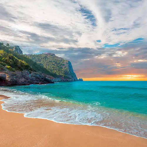 Ein breiter Sandstrand und türkisfarbenes Wasser mit leichter Brandung. Links steigt eine mit Bäumen bewachsene, felsige Klippe auf. Der Horizont glüht orange und gelb unter einem bewölkten Himmel bei Sonnenuntergang.