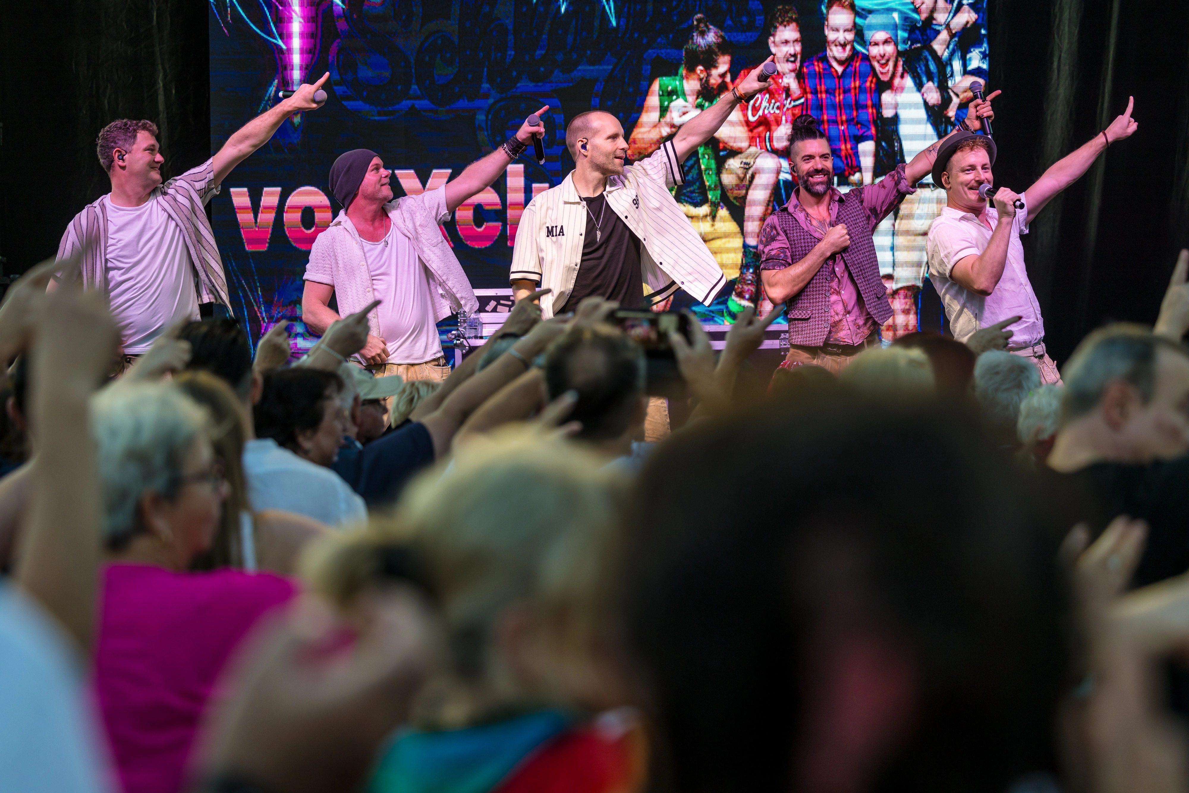 Eine Schlagerband steht auf der Bühne mit Sänger, Keyboarder und Gitarrist. Bunte Lichter beleuchten die Szene, während das Publikum tanzt und mitsingt. Schlagerband spielt live auf der Bühne, während Gäste tanzen und mitsingen.