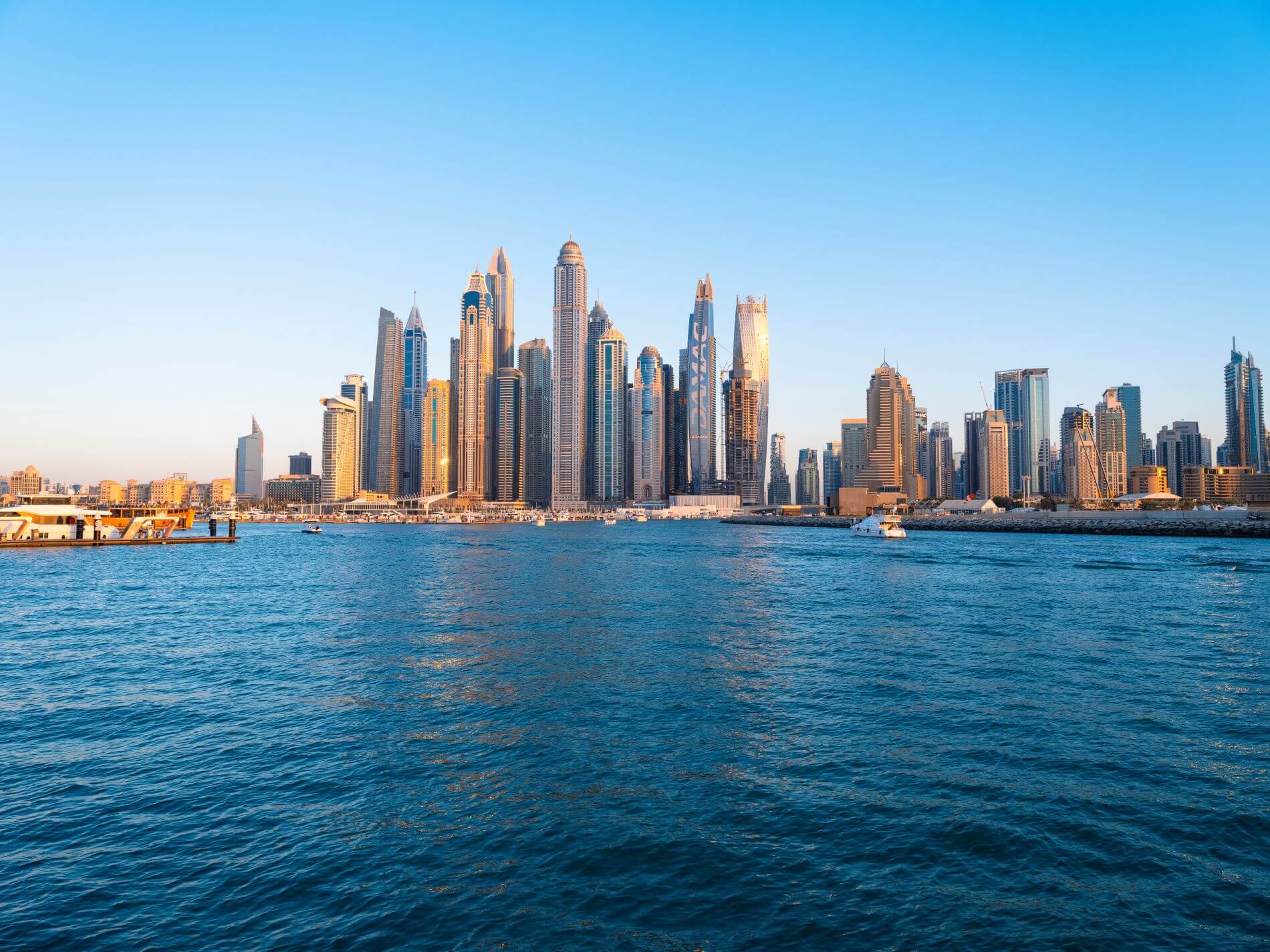 Die neue Marina in Dubai mit ihren Wolkenkratzer in der Dämmerung unter goldgelber Sonne an einem schönen, wolkenlosen Tag. In der Marina liegen zahlreiche kleine Boote vor Anker.
