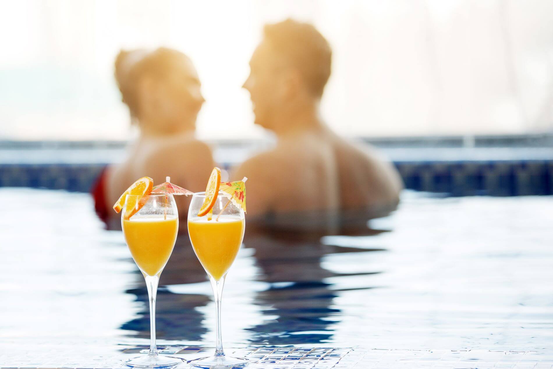 Zwei Gläser Orangensaft mit Scheiben stehen am Pool. Im Hintergrund steht ein Paar im Pool und unterhält sich.