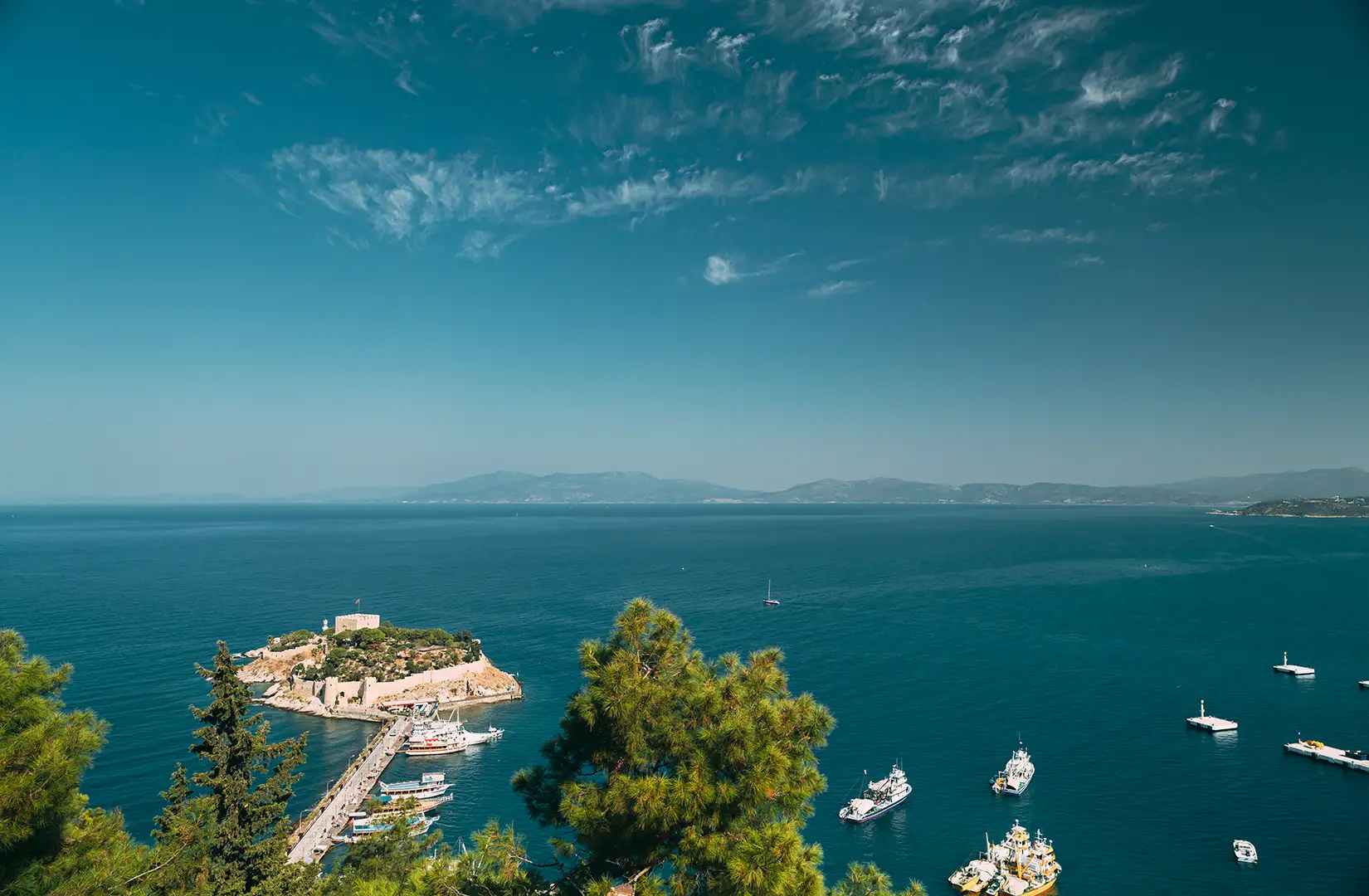Weitblick über das tiefblaue Ägäische Meer. Im Vordergrund liegt eine kleine, bewaldete Insel mit einer Burgruine und einem Pier, verbunden durch eine Fußgängerbrücke zum Festland. Yachten und Boote ankern im klaren Wasser.