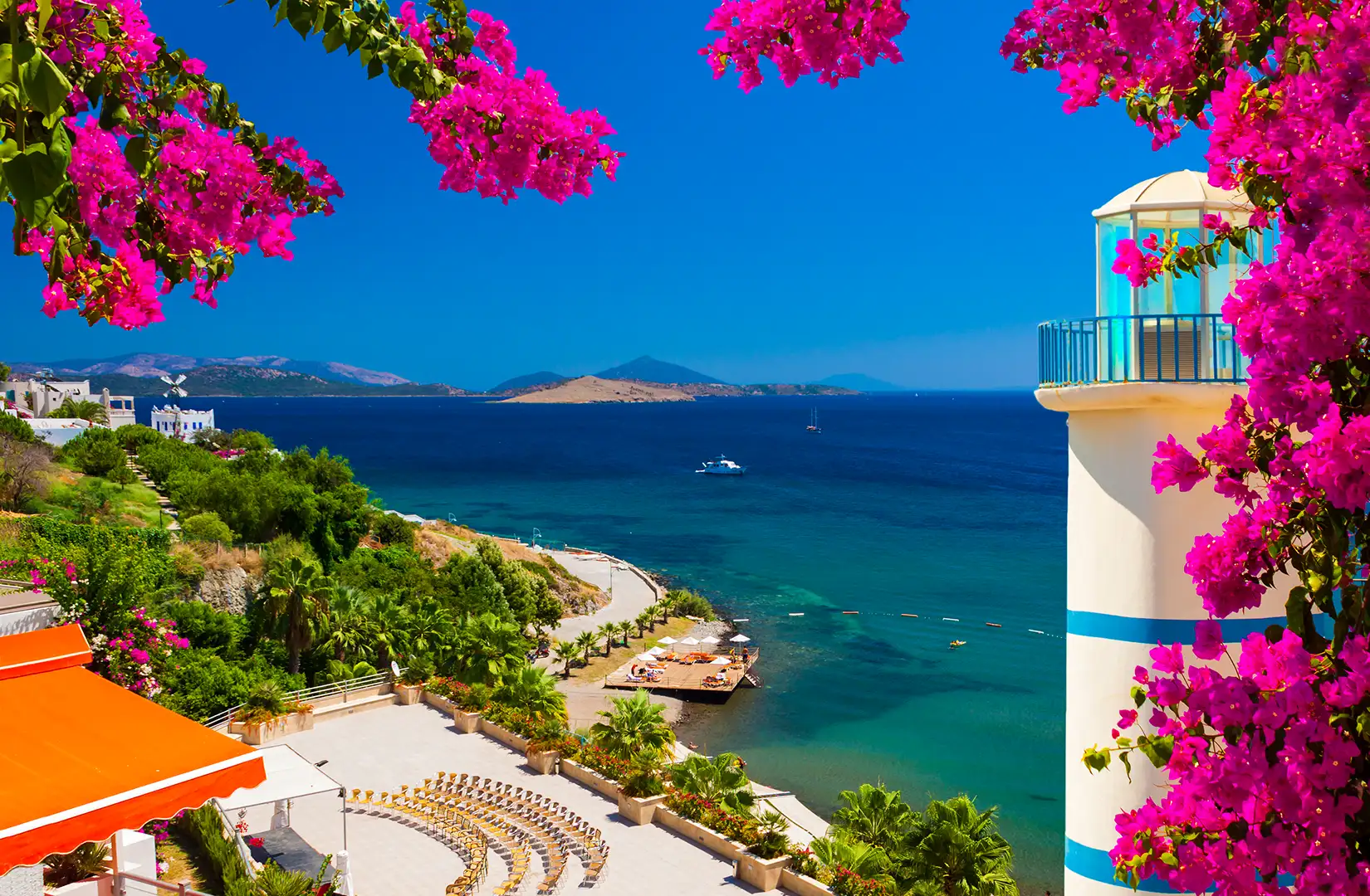 Ein sonniger Panoramablick über das tiefblaue Meer der Ägäis in Bodrum, eingerahmt von leuchtend pinker Bougainvillea. Im Vordergrund sind ein weißer Leuchtturm, eine Sonnenterrasse und üppige grüne Vegetation zu sehen.
