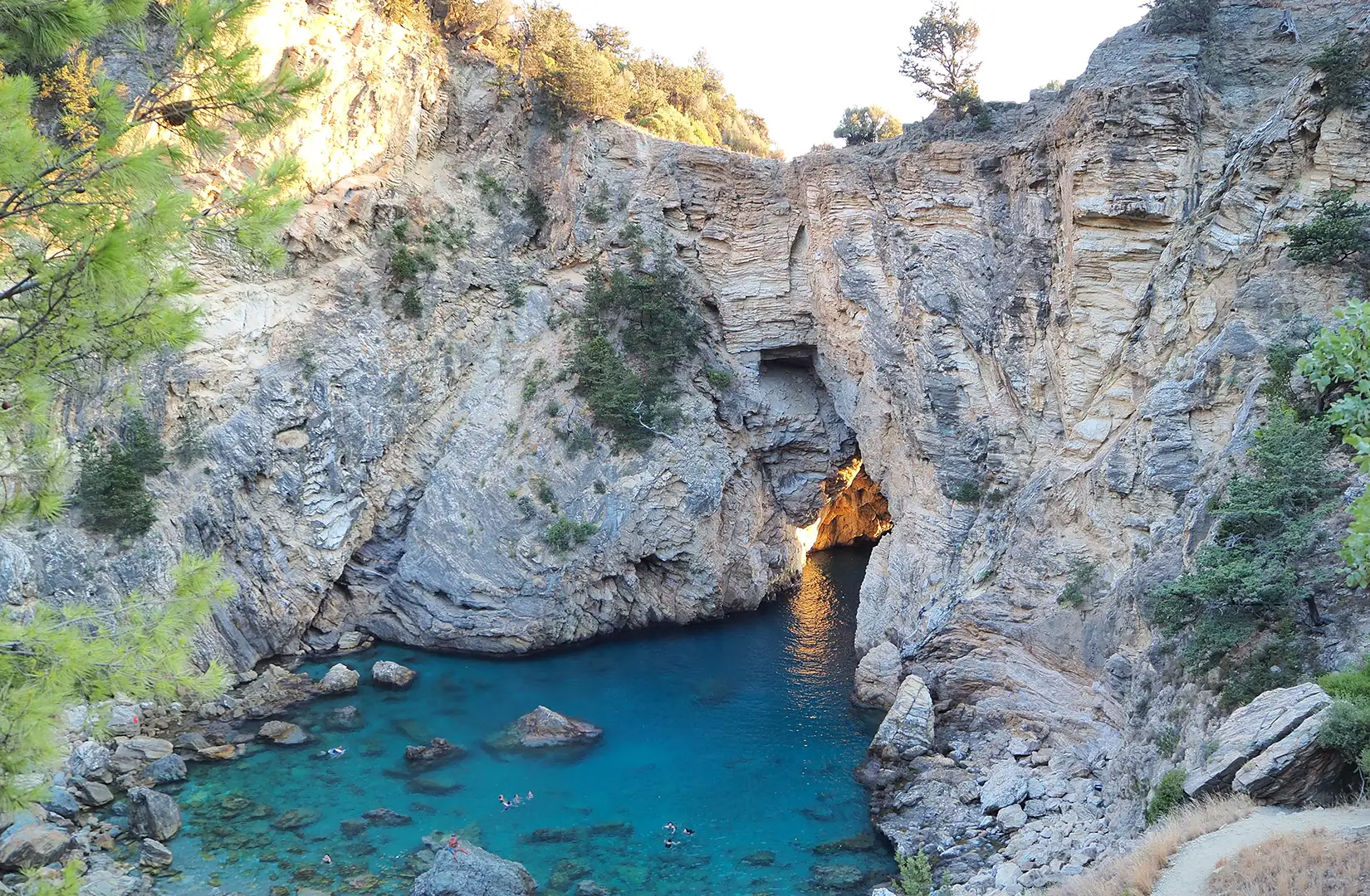 Eine enge, türkisfarbene Meeresbucht in Antalya, umgeben von hohen, zerklüfteten Felswänden. Das leuchtend blaue Wasser führt zu einer dunklen Höhle, die vom Sonnenlicht am Eingang goldgelb beleuchtet wird.