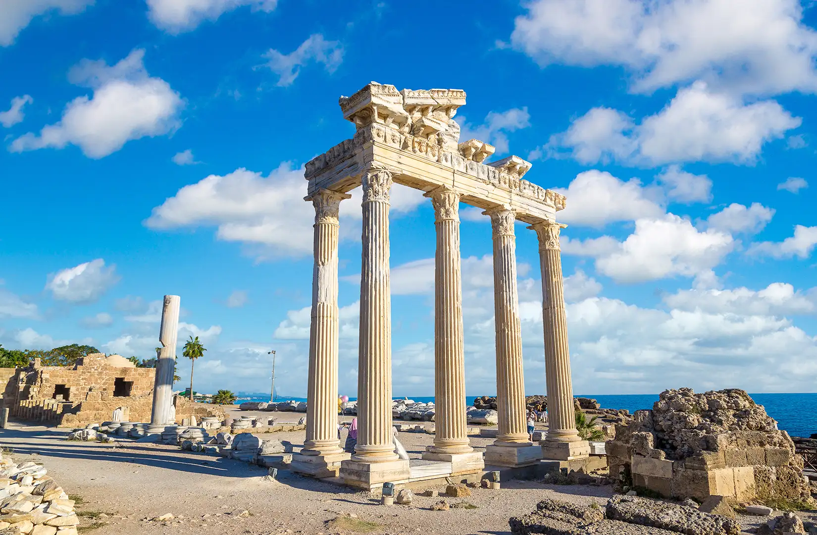 Die majestätischen Überreste des Apollo-Tempels in Side mit vier hohen, cremefarbenen korinthischen Säulen stehen am Rande des Meeres. Ein strahlend blauer Himmel mit weißen Wolken bildet den Hintergrund der antiken Stätte.